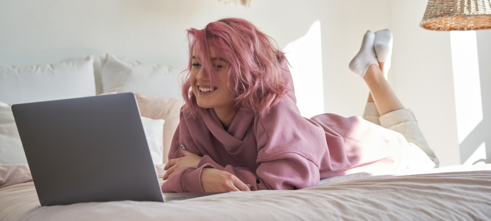 Teenie Mädchen mit rosa Haaren liegt im Bett und schaut auf den Laptop