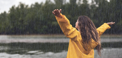 Frau steht mit gelber Regenjacke und erhobenen Armen im Regen