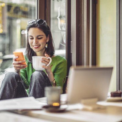 Junge Frau sitzt mit Handy und Laptop im Café.